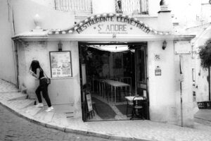 black and white photograph of street scene in Lisbon outside cafe with figure on the left climbing steps that are part of the sidewalk.