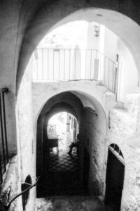 black and white photograph of monastery at Wadi Qelt, exterior is white painted stone with arched passage and doorways. A figure is seated on the right at the exit of passageway framed by bright light in the background