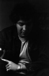 Black and white photograph of a figure in shadow looking down. Hand is visible at bottom with index finger pointing to left. Figure is wearing a grey/white turtleneck and dark jacket. Figure has a silver bracelet around wrist.