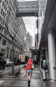 City scene photograph in mostly black and white with a female figure in color wearing a red hoodie, jean shorts and boots walking away on wet sidewalk